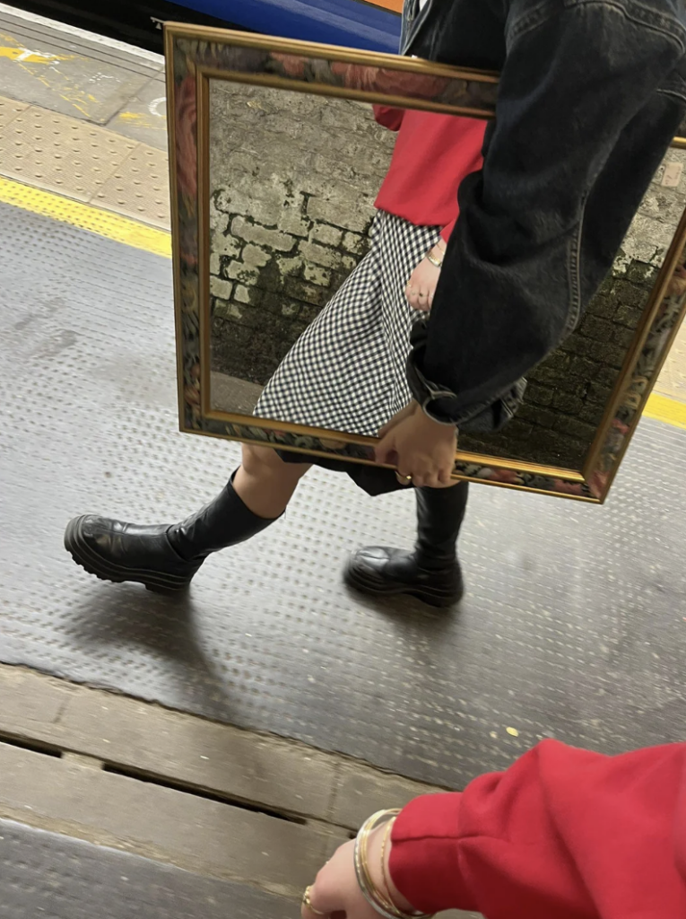 A person walking on a train platform carrying a large, rectangular mirror with an ornate frame. The person is wearing a red top, black denim jacket, a black and white checkered skirt, and black boots. Another person's arm, wearing a red sleeve, is visible in the foreground.