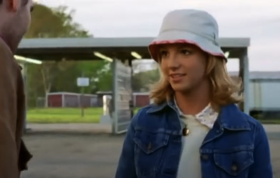 A person with shoulder-length blonde hair wears a white bucket hat and a denim jacket over a light-colored top. They are outdoors near a structure with a covered area. Trees and a building are visible in the background.