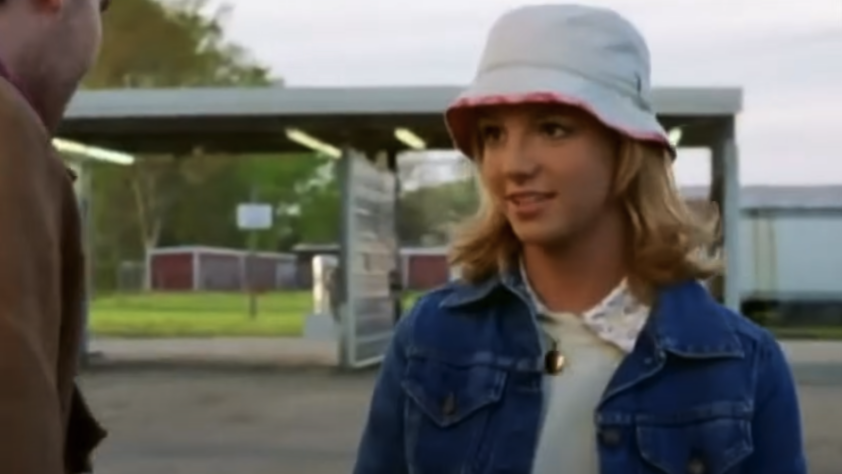 A person with shoulder-length blonde hair wears a white bucket hat and a denim jacket over a light-colored top. They are outdoors near a structure with a covered area. Trees and a building are visible in the background.