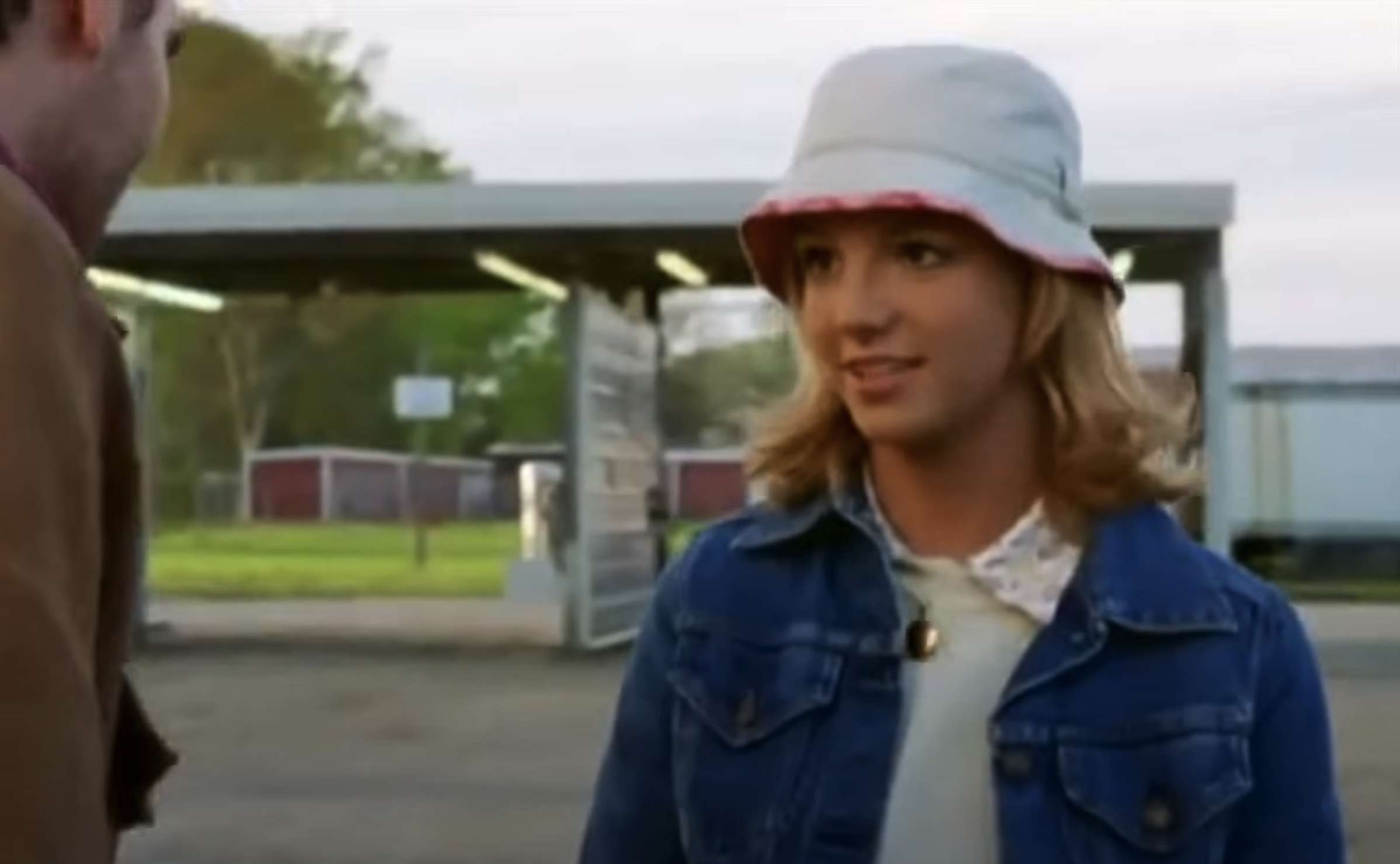 A person with shoulder-length blonde hair wears a white bucket hat and a denim jacket over a light-colored top. They are outdoors near a structure with a covered area. Trees and a building are visible in the background.