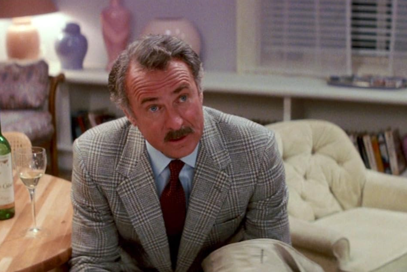 A man with graying hair and a mustache, wearing a gray checkered suit, blue shirt, and red tie sits in a living room. A glass of wine and a bottle are on the table nearby. The background has a bookshelf, a cream armchair, and decorative items.