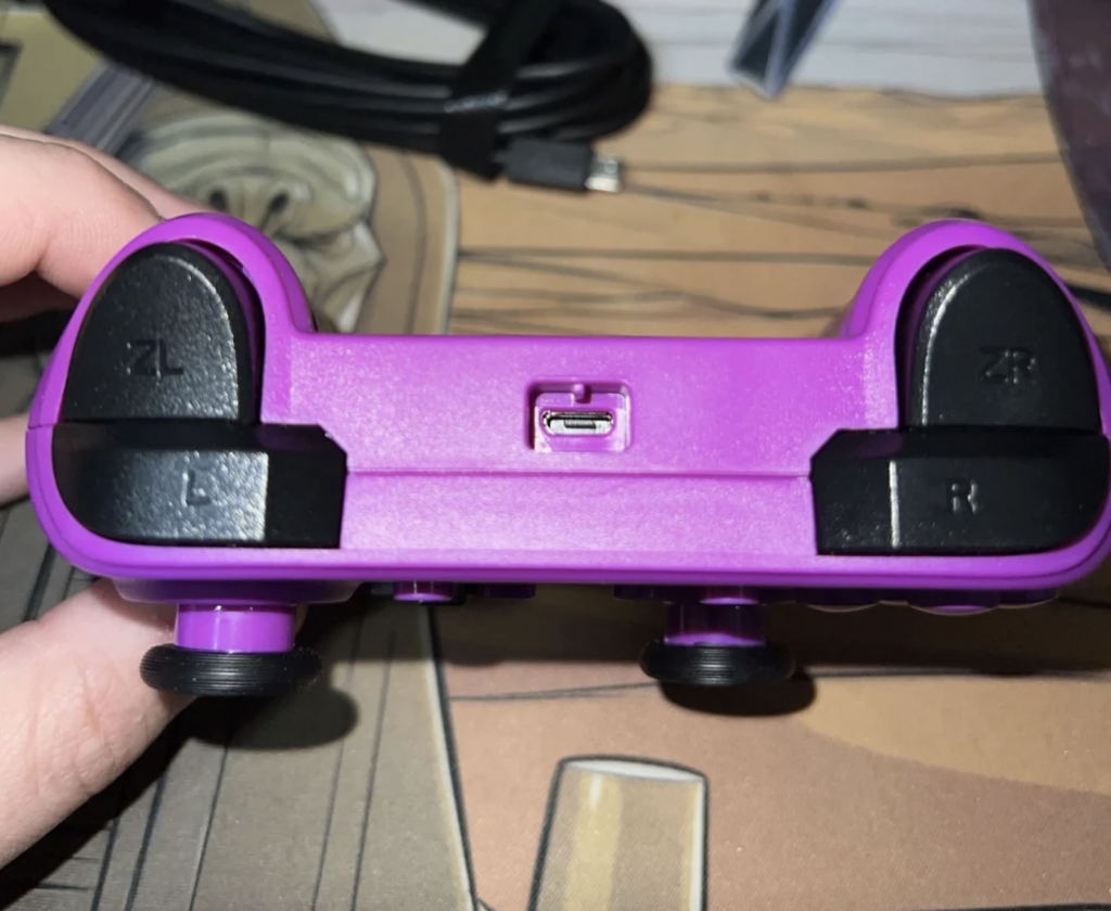 A person holding a purple gaming controller upside down, showing the trigger buttons labeled ZL, L, ZR, and R. In the middle, there is a small USB charging port. A cable and some background items are also partially visible.