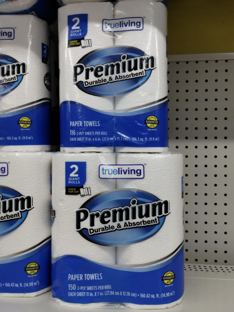 Packages of true living Premium paper towels on a store shelf. The packaging highlights that each package contains two giant rolls, and the towels are described as durable and absorbent. The differences in the packs on display are in the number of sheets per roll.