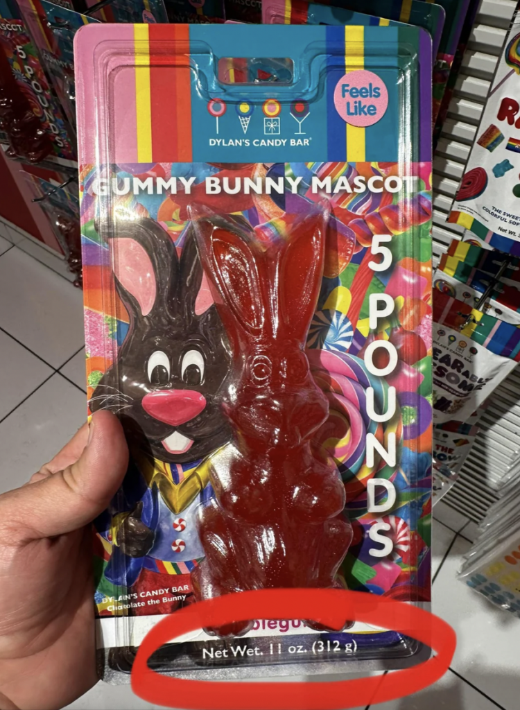 A person holds a package of a candy shaped like a bunny. The label reads "Gummy Bunny Mascot," "5 Pounds," and "Feels Like." The net weight displayed at the bottom is 11 oz (312 g). The background includes colorful candy and text.