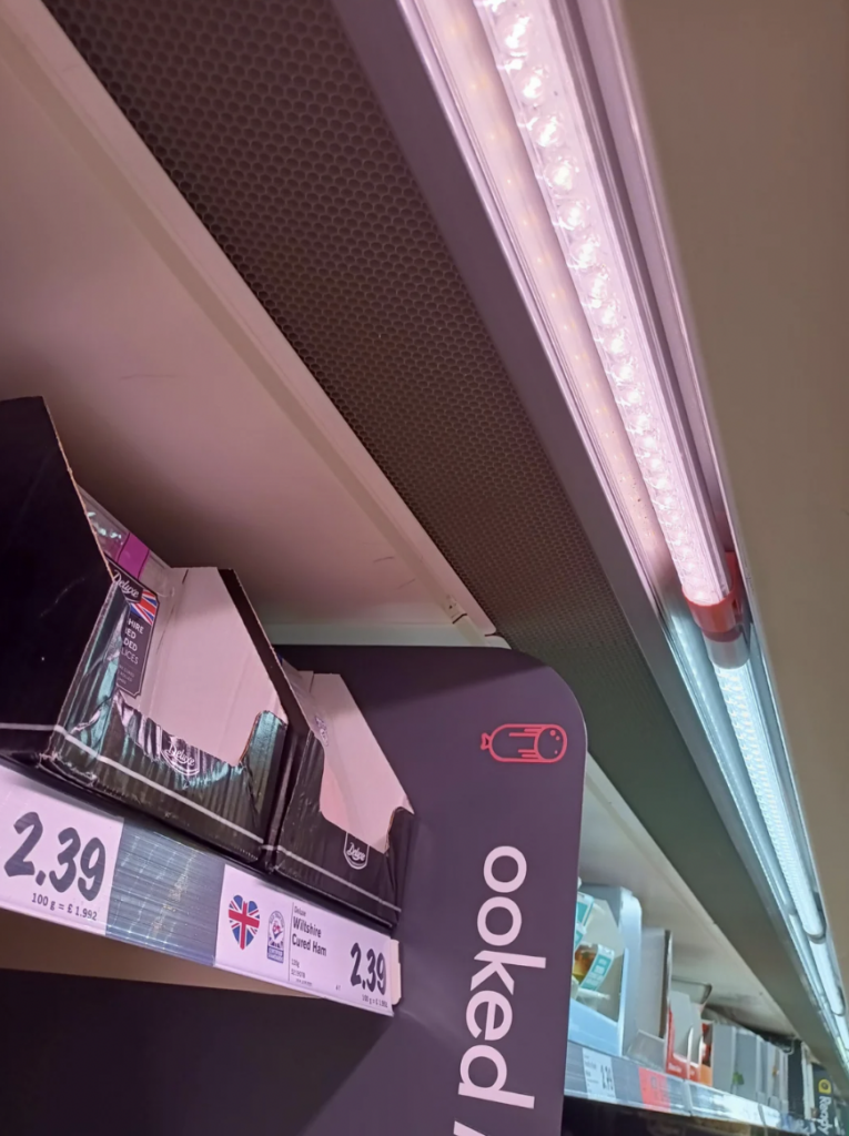 A supermarket shelf displays packaged goods with a price tag of £2.39. Above the shelf are two fluorescent lights, one fully lit and the other partially visible. The edge of the display features cut-off text reading "cooked.