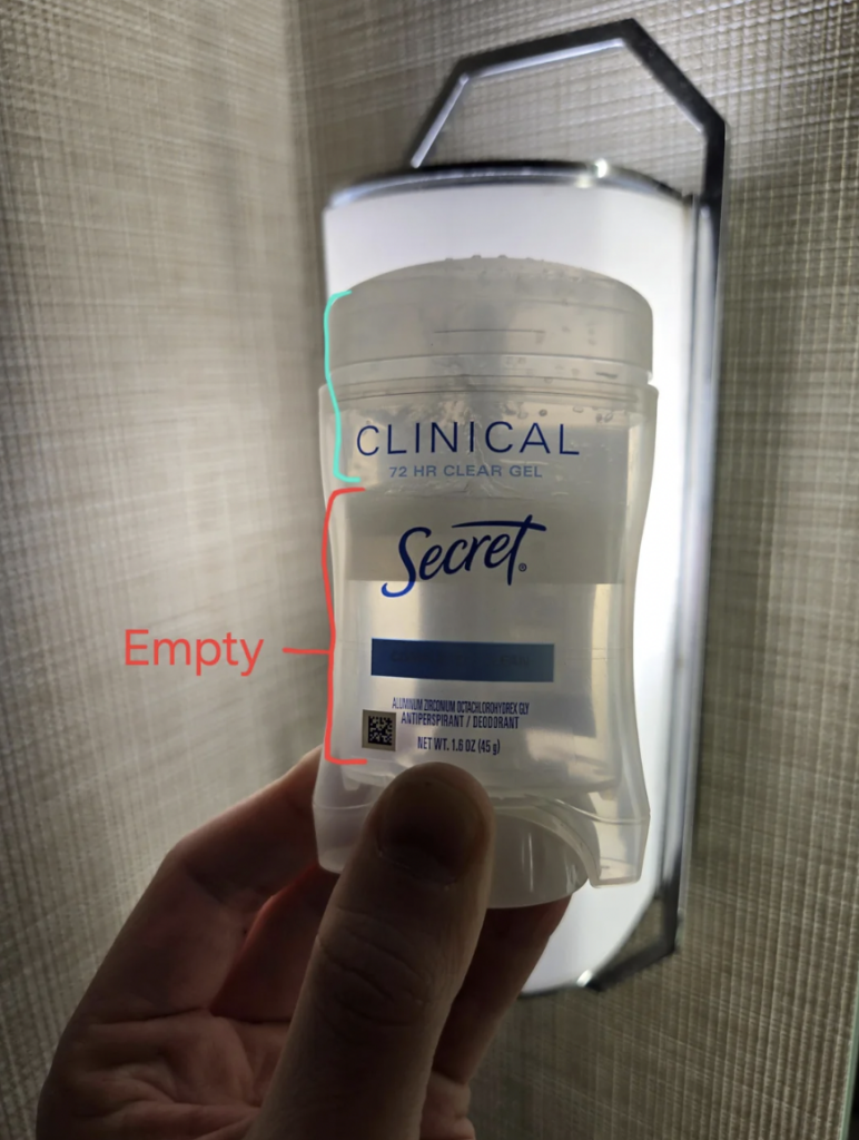 A person holds an empty container of Secret Clinical 72 HR Clear Gel antiperspirant deodorant against a lightly textured background. The word "Empty" is annotated on the image with an arrow pointing to the empty portion of the container.