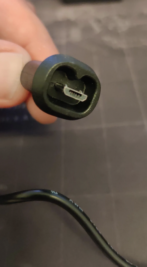 A close-up image of a hand holding a small, black power connector. The connector is oval-shaped with a small, metal pin inside. The background is out of focus, revealing a grid-patterned surface.