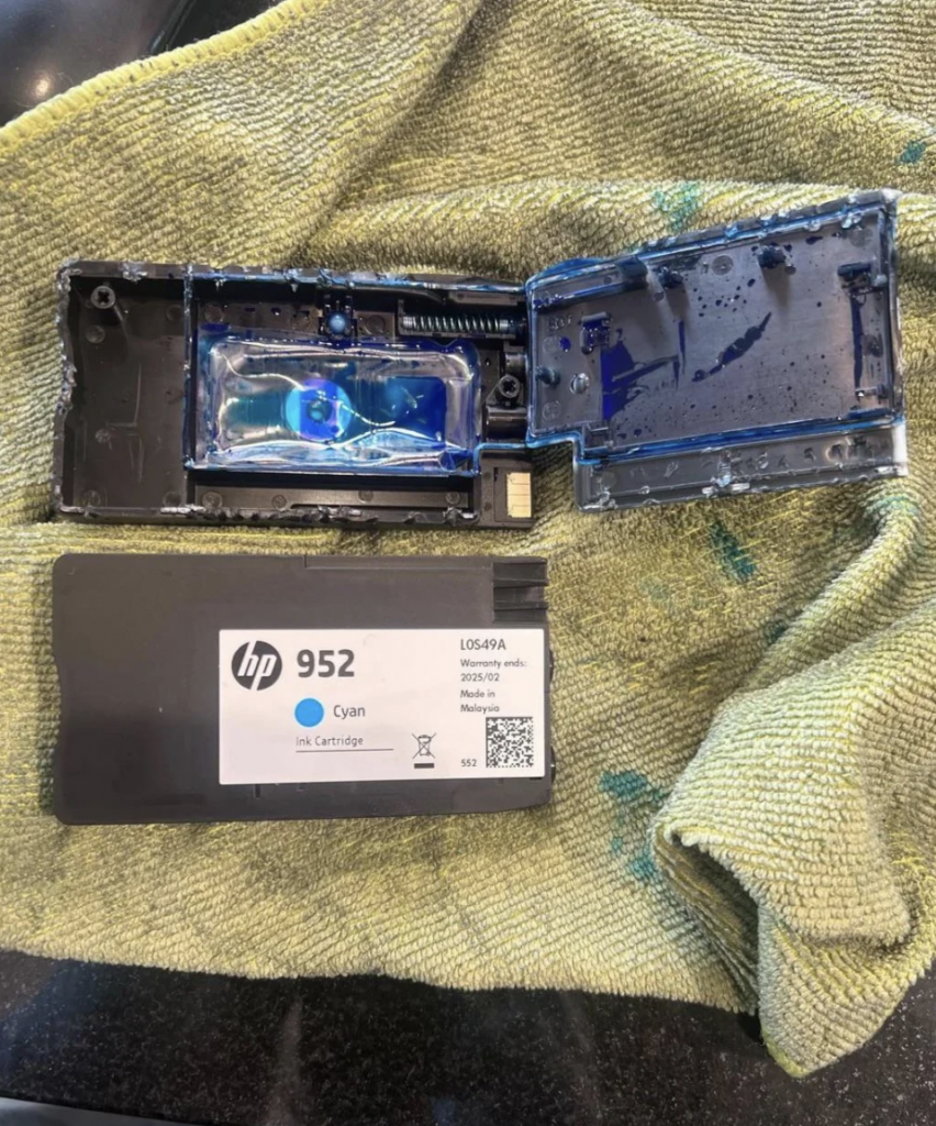 An opened HP 952 cyan ink cartridge is placed on a green towel. The cartridge's components and spilled blue ink are visible, indicating it has been tampered with. The cartridge label is clearly visible, displaying the model number and color.