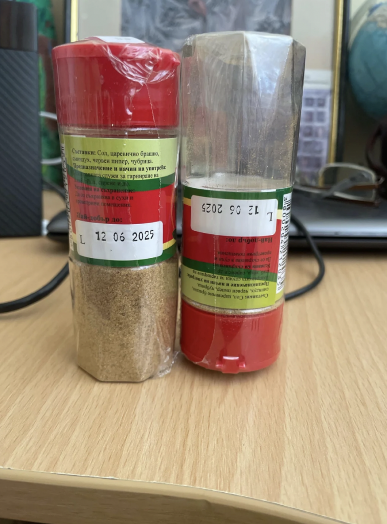 Two spice containers with red caps sit side by side on a wooden surface. Both containers have labels with text and a printed expiration date of "12 06 2025." The contents in one bottle are visible, showing a brown spice, while the other bottle is partially wrapped in plastic.