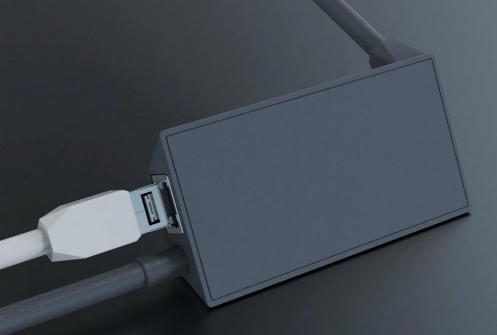 A close-up of a grey adapter box on a dark surface, with a white USB-C cable plugged into it on the left side and a grey cable connected on the right side. The adapter has a minimalistic design with a smooth, featureless surface.