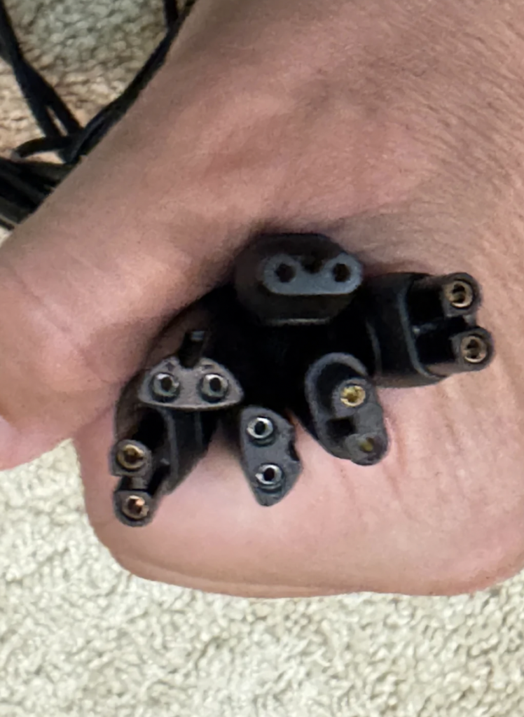 A hand holding an assortment of black power cable connectors, each with three prong holes and varying shapes. The background appears to be a textured, off-white surface.