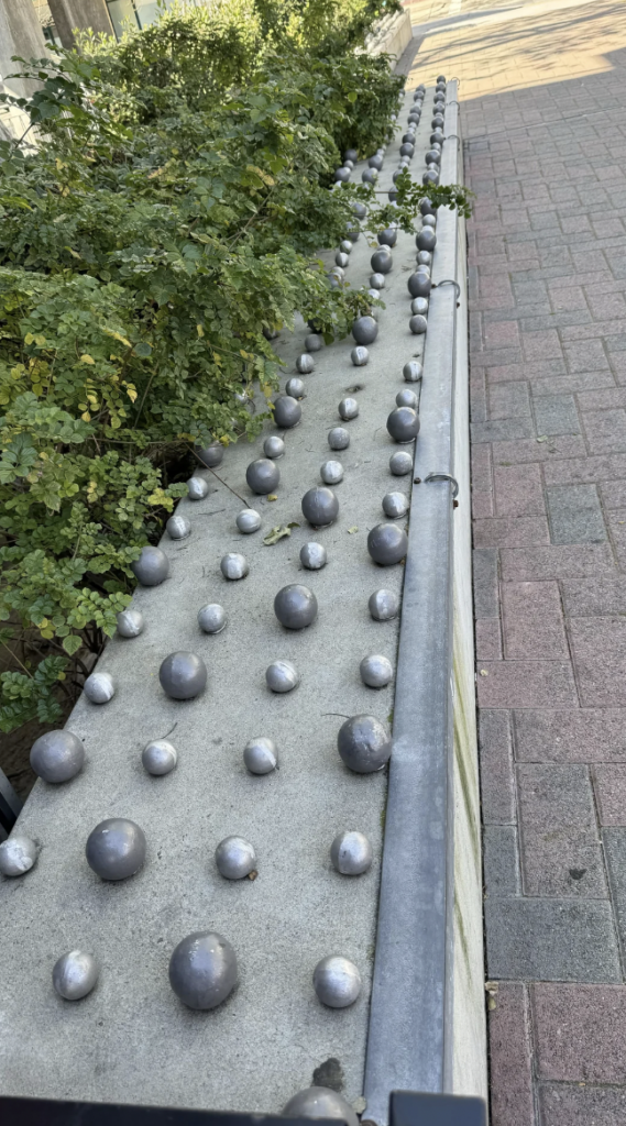 Concrete planter with numerous metallic spheres of varying sizes affixed to its surface, surrounded by lush green plants and bordered by a red and grey brick pathway.