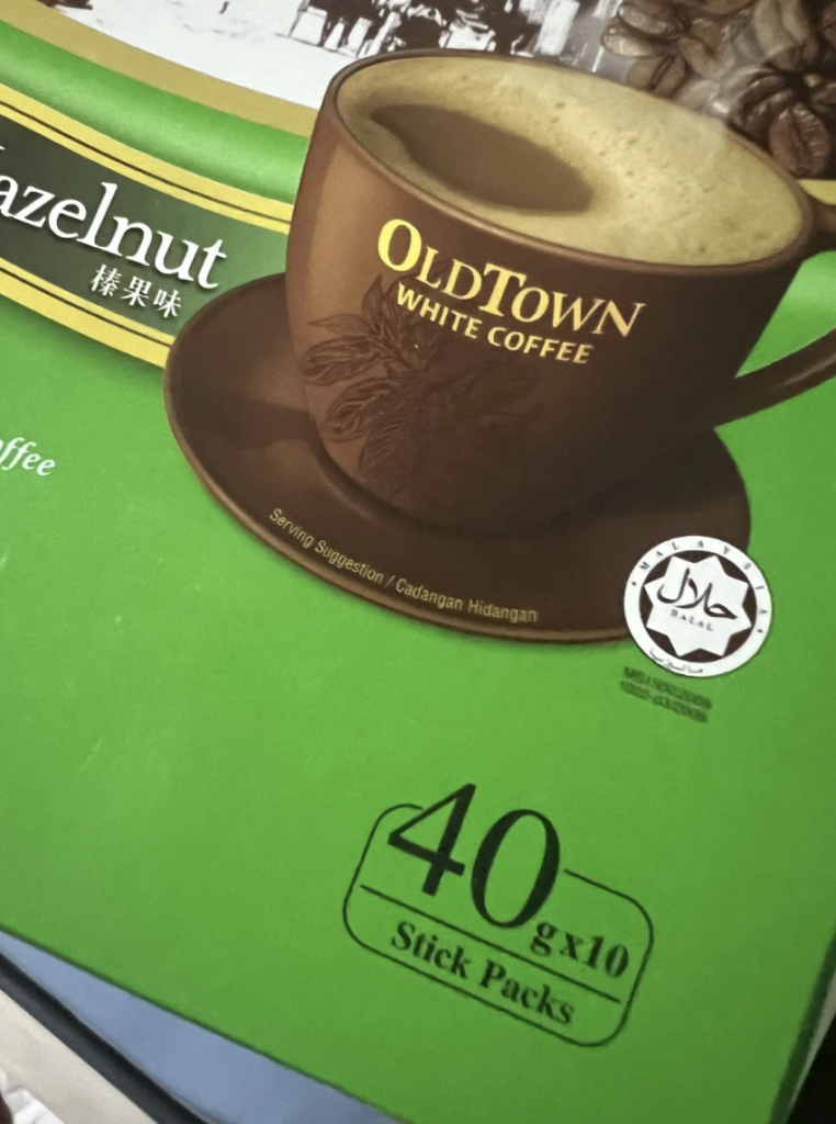 A green package of OldTown White Coffee Hazelnut flavor with a picture of a coffee cup filled with creamy coffee. The text "40g x 10 Stick Packs" is displayed, and the package also features a Halal certification symbol.