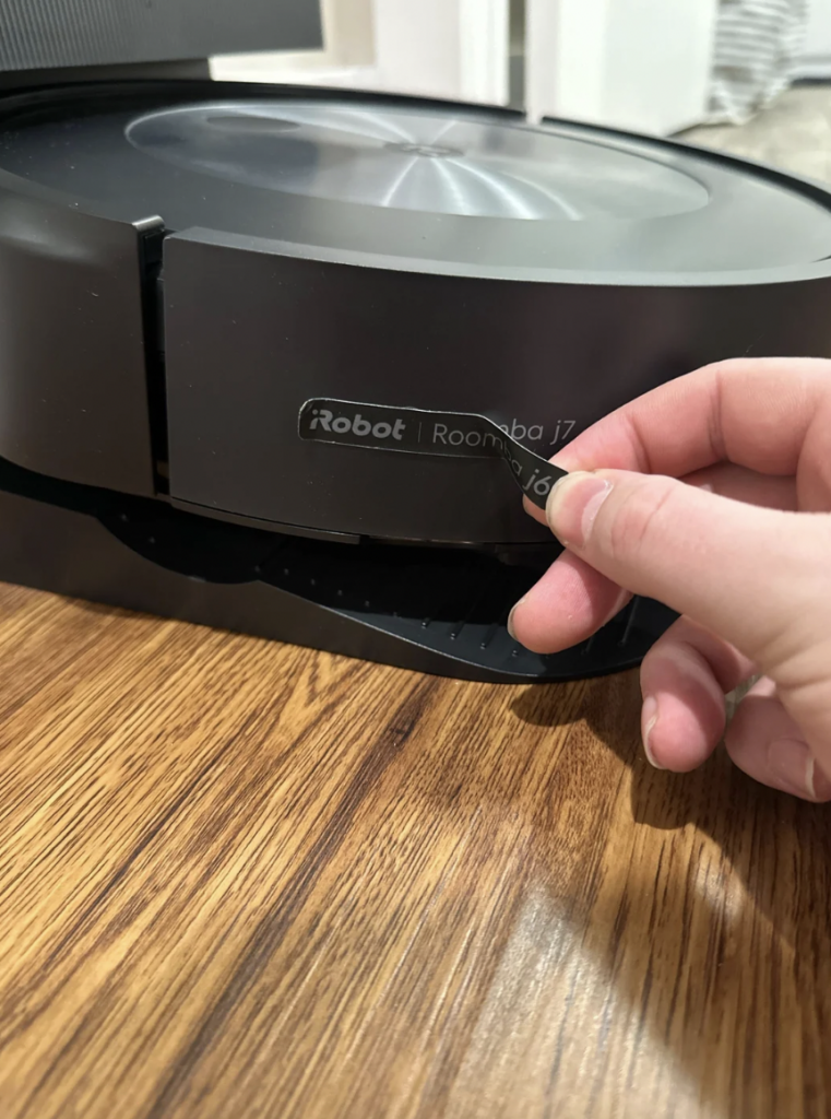 A close-up of a hand peeling off a protective sticker from a black iRobot Roomba j7+ robot vacuum positioned on a wooden floor. The hand is partially covering the text on the sticker while removing it.