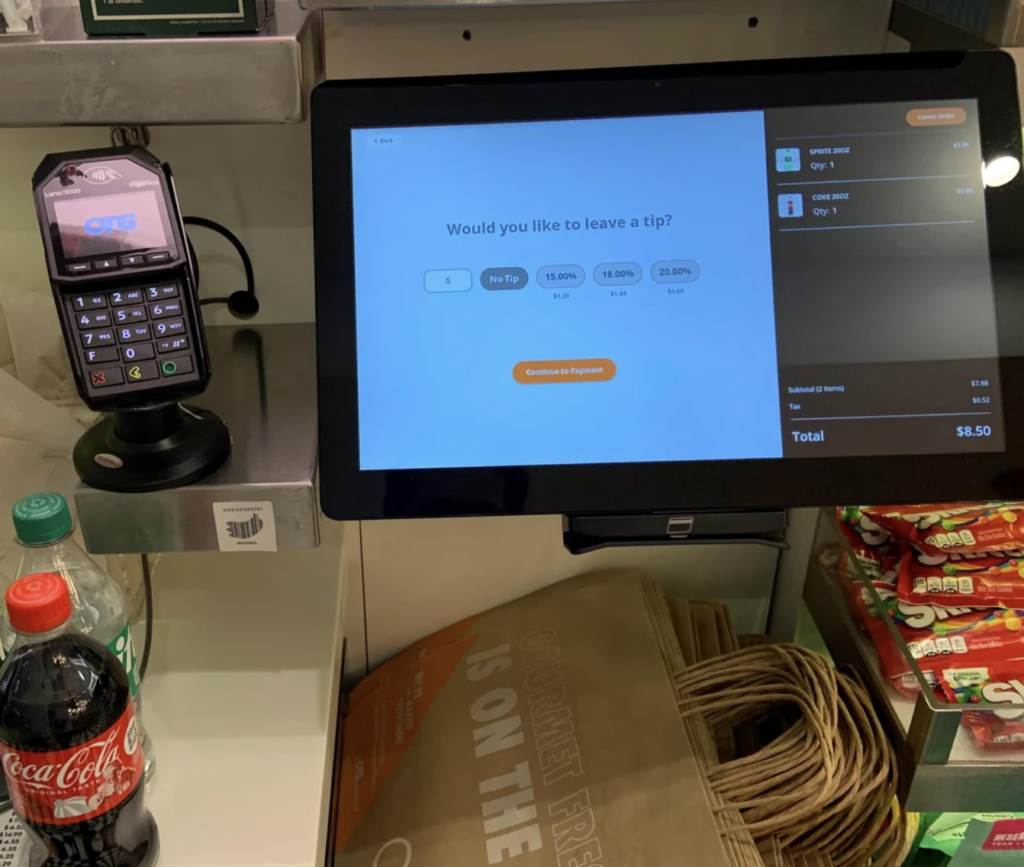 A touchscreen display at a checkout counter shows tip options and a payment summary for $8.50. Below the screen, a large paper bag sits on a shelf with bottled beverages and candy nearby. To the left, a card reader is mounted on the counter.