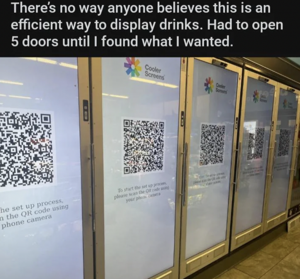 A row of refrigerator doors in a store, each featuring large QR codes on digital screens with instructions for a set-up process. A sign on top reads, "There's no way anyone believes this is an efficient way to display drinks. Had to open 5 doors until I found what I wanted.