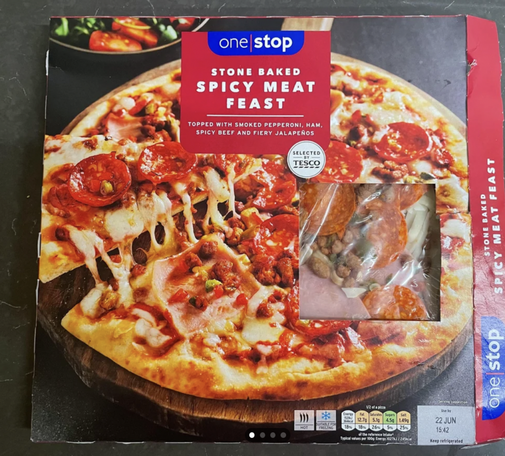 Box of One Stop "Stone Baked Spicy Meat Feast" pizza with a picture of a cooked pizza topped with smoked pepperoni, ham, spicy beef, and jalapenos. The box has a clear window showing the uncooked pizza inside. Tesco logo is visible.