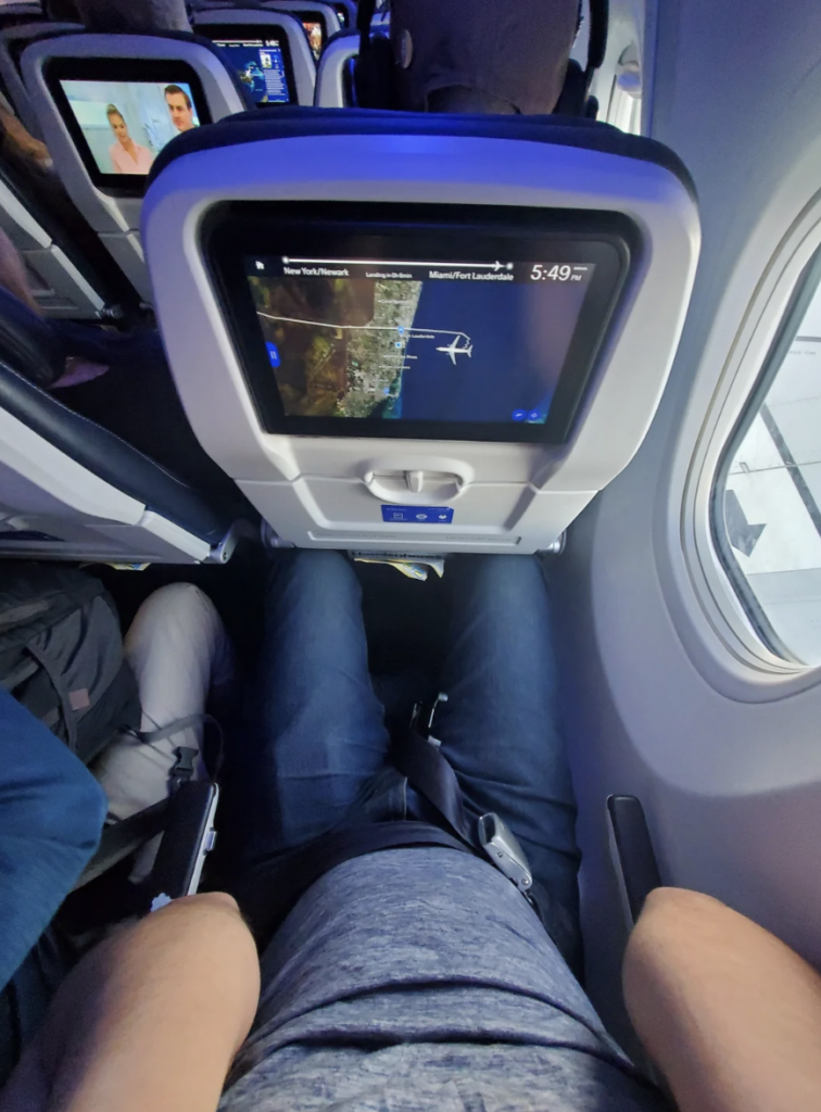 A person is seated on an airplane, viewing an in-flight entertainment screen showing a map. The time displayed is 5:49. The passenger's legs and a glimpse of the plane window are visible. The person is wearing jeans and a gray shirt.