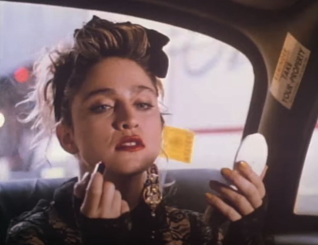 A woman with blonde hair, adorned with a black bow, is sitting in a car and applying lipstick while looking into a small mirror. She is wearing a black lace outfit and large earrings, with an urban setting visible through the window behind her.