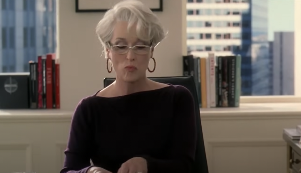 A person with short, styled gray hair and glasses sits at a desk in an office, wearing a dark blouse and hoop earrings. They appear focused, looking down at what they are doing. Shelves with books and a large window with a cityscape view are in the background.