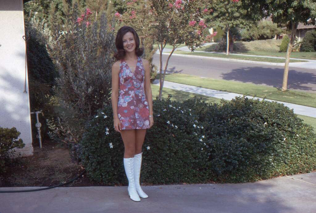 A woman with dark hair stands outside in front of a bush and trees on a sunny day. She is wearing a sleeveless, floral-patterned dress and white knee-high boots. A quiet suburban street is visible in the background.