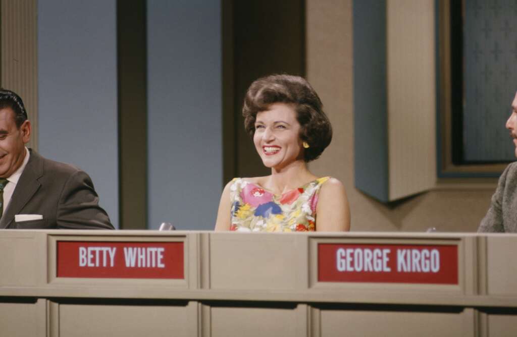 A woman with short brown hair in a colorful floral dress sits smiling behind a desk on a game show set. To her left, a man in a suit is partially visible. Nameplates in front of them read "BETTY WHITE" and "GEORGE KIRGO.