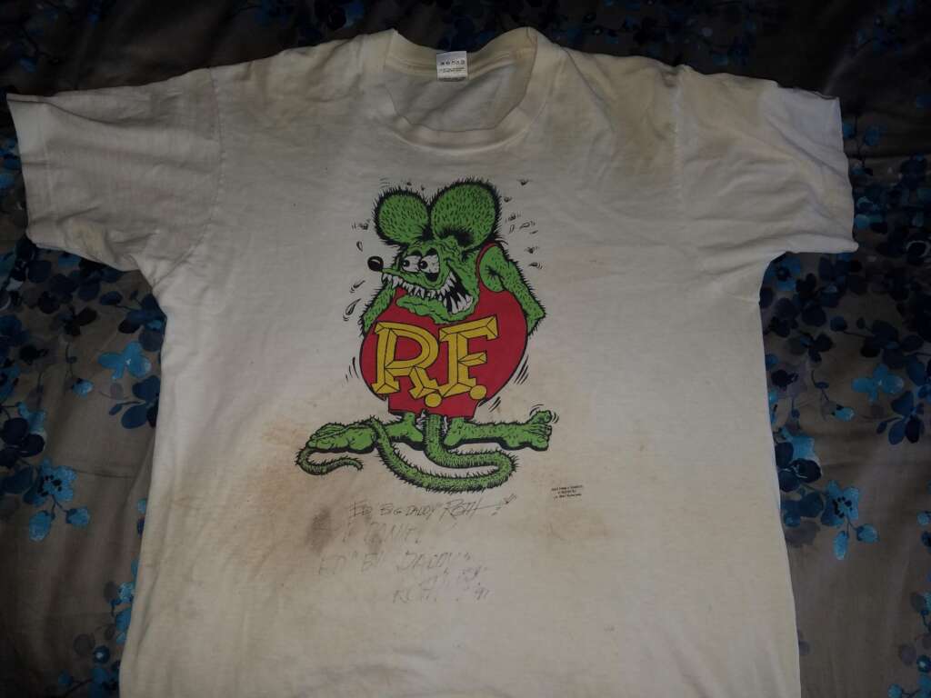 A white t-shirt with an illustration of a green, anthropomorphic rat holding a red heart with the letters "RF" in yellow. The shirt is slightly stained and worn, with blue floral bedding in the background.