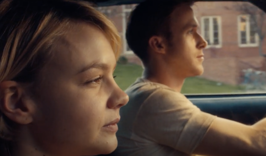 A woman and a man sit in a car, both focused ahead. The woman is nearer to the camera, showing a thoughtful expression. The man is driving, his profile visible. The background shows a brick building through the car windows.