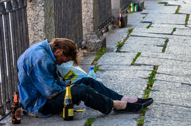 A man is sitting on a sidewalk, slumped forward with his head down. He is wearing a denim jacket and dark pants. Around him are several bottles and cans, and he appears to be surrounded by litter. The setting is along a stone path with a metal railing.