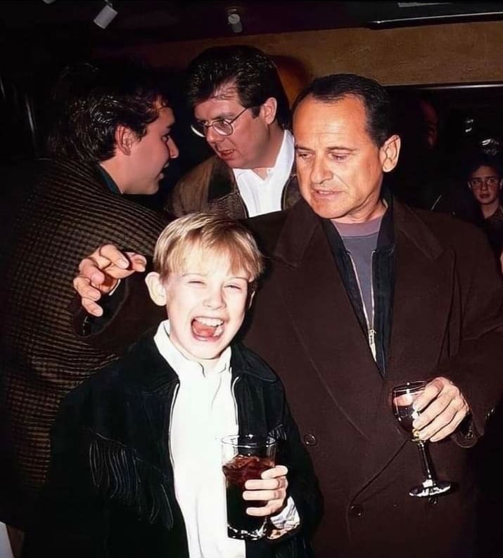 A young boy, holding a drink, is laughing joyfully while a man next to him, holding a wine glass, looks down at him with a smile. Two other men in the background are engaged in conversation. They appear to be in a dimly lit room, possibly at a social event.