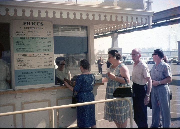 A group of people stands in line at an outdoor ticket booth. One person is purchasing tickets from the clerk behind the booth window. A price board lists different admission and ticket book options for adults and children. A bridge structure is visible in the background.