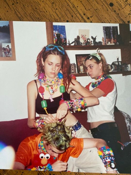 Three individuals with colorful, eccentric attire are seen in a room. One stands behind a seated person, braiding their green hair while another stands beside them. All three wear an abundance of bright beaded jewelry and vibrant clothing.背景には棚に小さな装飾品が並んでいます。