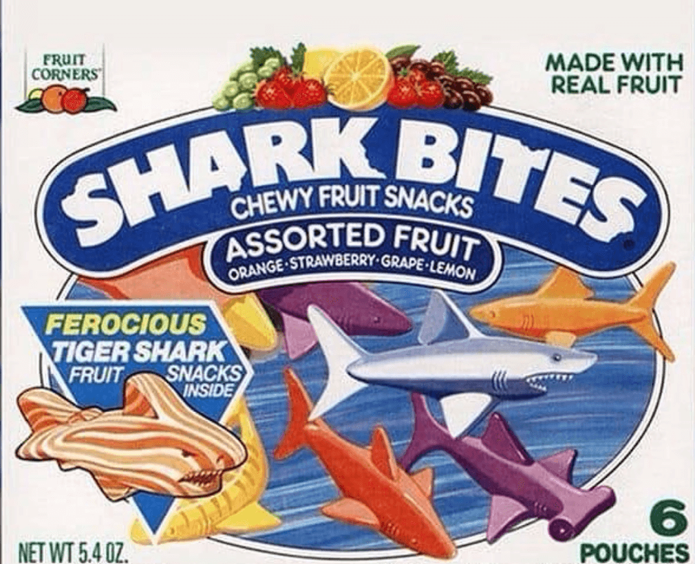 The image shows a box of Shark Bites chewy fruit snacks featuring "assorted fruit" flavors: orange, strawberry, grape, and lemon. The box contains 6 pouches. The packaging highlights a "ferocious tiger shark" fruit snack and mentions "made with real fruit.