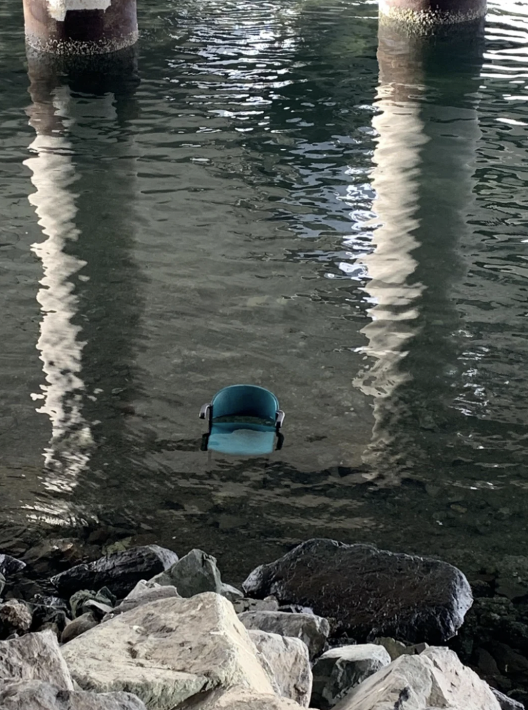 Partially submerged in water under a bridge, a blue plastic chair is surrounded by large rocks. Three pillars of the bridge are reflected in the water, creating a symmetrical pattern. The scene evokes an unusual and serene atmosphere.