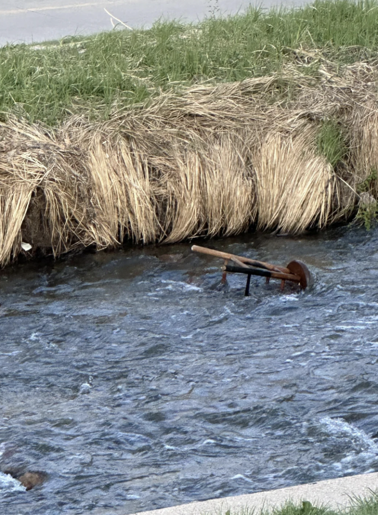 A rusted, overturned bike lies partially submerged in a flowing stream. The stream is bordered by dry, tall grasses on one side and a concrete edge on the other. The scene suggests neglect and abandonment.