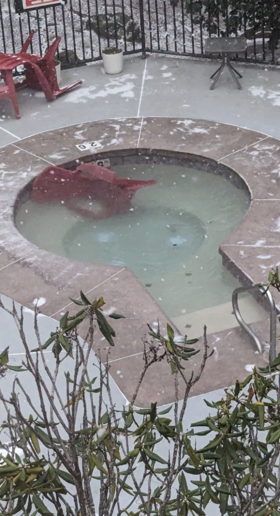 A jacuzzi with partially submerged red patio chairs in icy water. Snow lightly covers the surrounding concrete and plants around the jacuzzi, suggesting a cold and snowy environment. The jacuzzi's steps and metal handrail are also slightly snow-covered.