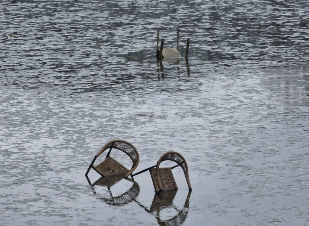 Two overturned wicker chairs partially submerged in floodwater. In the background, two wooden poles pierce the water's surface. The gray water reflects the overcast sky, creating a somber and tranquil scene.