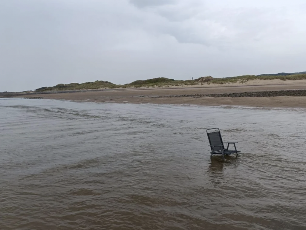 A solitary folding chair is partially submerged in shallow water near the shore on a cloudy day. The sandy beach and grassy dunes are visible in the background under an overcast sky.