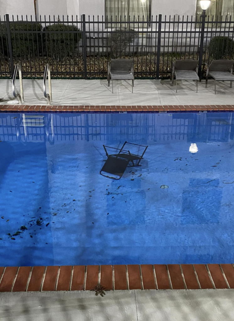 A nighttime view of an outdoor swimming pool with a metal chair submerged underwater. The pool is surrounded by brick coping, a tall black iron fence, and several empty lounge chairs. The water shows reflections of nearby lights.