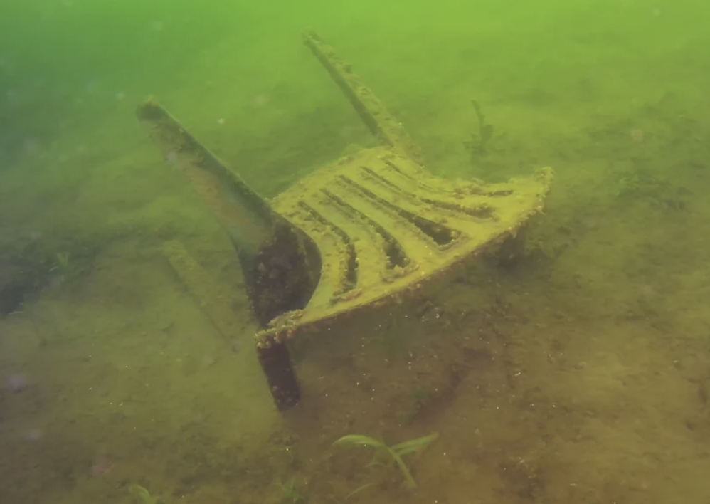 A plastic chair lies partially buried and upside down on the muddy bottom of a greenish, murky body of water. Algae and small aquatic plants are growing on the surface and surrounding areas. Bubbles and particles float in the water, creating a slightly cloudy environment.