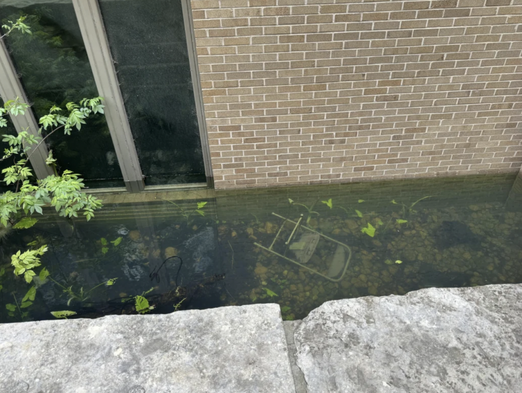 A brick building with a large window is partially submerged in water, indicating severe flooding. A chair and debris are visible underwater near the building, and some green plants float on the water's surface. Stone steps lead to the water's edge.