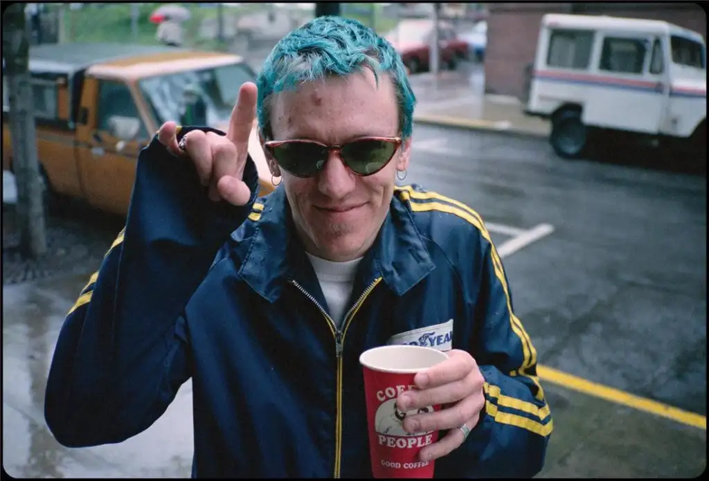 A person with short, blue hair and wearing sunglasses is standing outdoors on a rainy day. They are dressed in a blue and yellow track jacket and holding a red coffee cup. The person is making a hand gesture with their left hand, and a postal service vehicle is in the background.