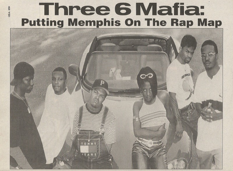 A black and white image of the hip-hop group Three 6 Mafia in front of a car. Six men are gathered, wearing casual 90s fashion like striped shirts, overalls, and jeans. A headline above reads, "Three 6 Mafia: Putting Memphis On The Rap Map.