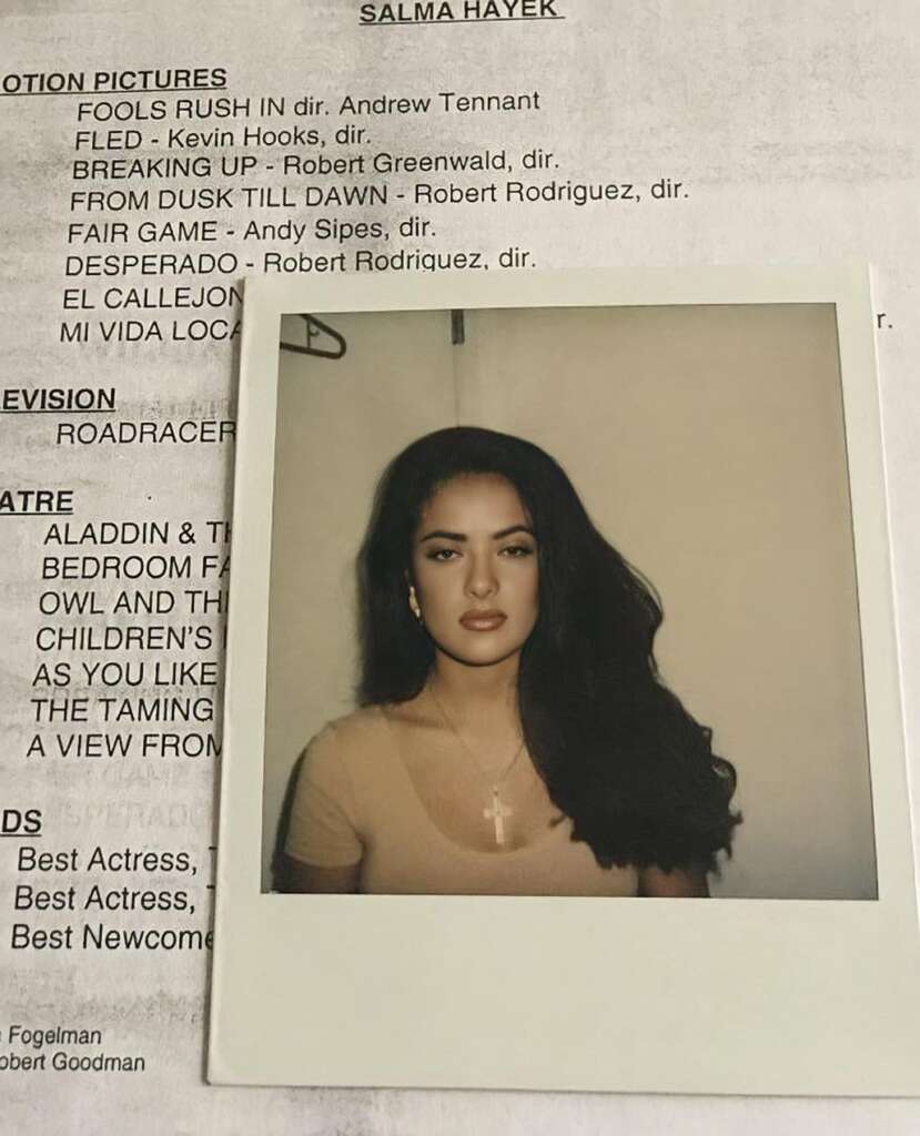 A Polaroid photo of a woman with long dark hair, wearing a brown top and a cross necklace, is placed on a paper listing movies and TV shows. Her expression is neutral, and she is facing the camera directly. The paper lists various titles and names.