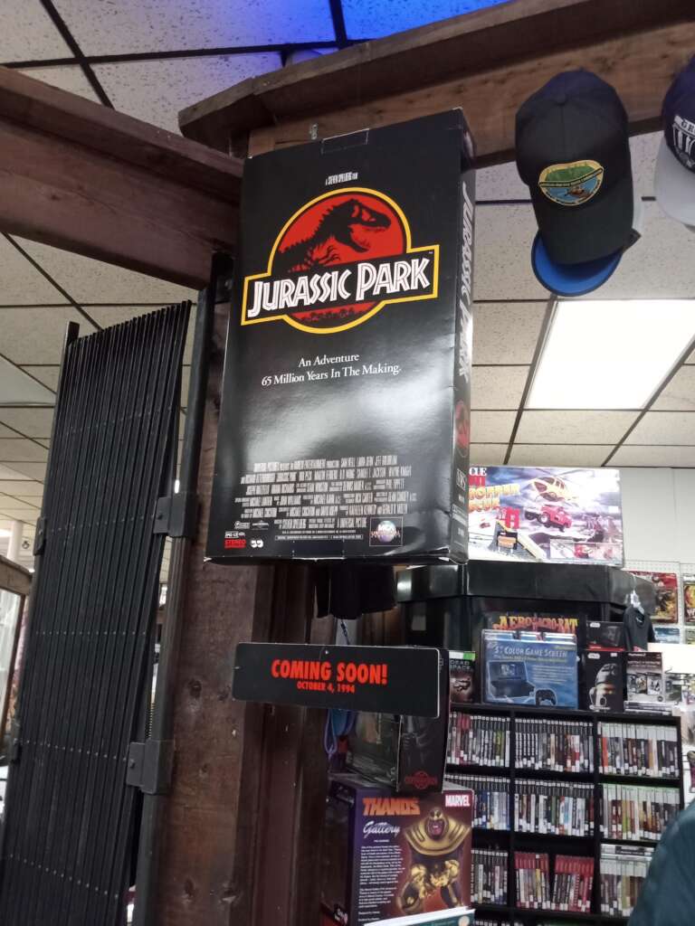 A store displays a "Jurassic Park" movie poster that reads "An Adventure 65 Million Years In The Making" with a release date of June 11, rated PG-13. Below the poster, a sign states "COMING SOON!" and the store has shelves stocked with various items and merchandise.