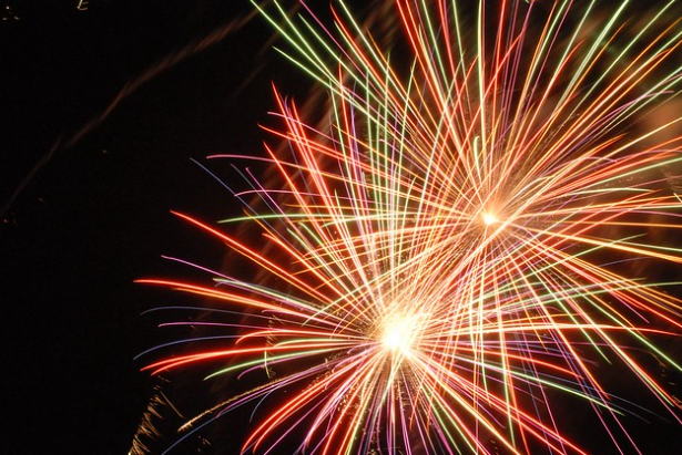 A burst of colorful fireworks illuminates the night sky, with streaks of yellow, orange, red, green, and purple radiating outward. The vibrant display creates a dazzling pattern of light against the dark backdrop.