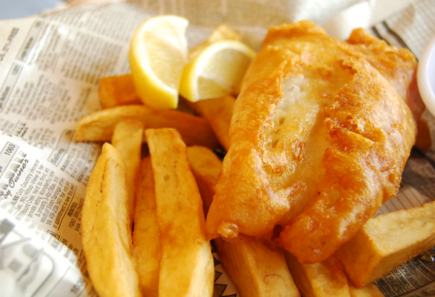 Close-up of a serving of fish and chips. The golden-brown fried fish sits on top of thick-cut fries, with lemon wedges on the side. The meal is presented on a piece of newspaper, adding a traditional touch.