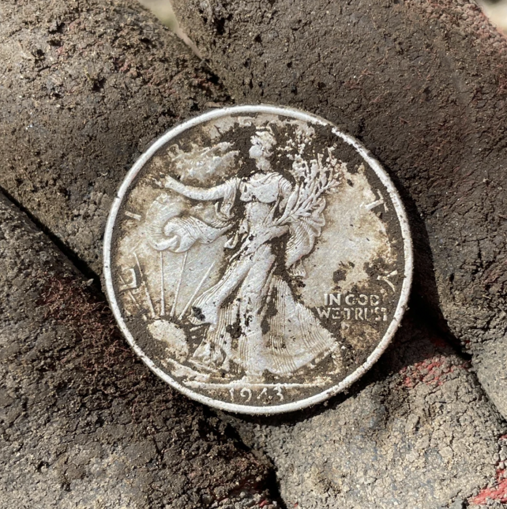 A worn 1943 Walking Liberty half dollar coin is held in a person's dusty, gloved hand. The details of Lady Liberty, along with the "IN GOD WE TRUST" inscription, are still partially visible despite the coin’s aged condition.