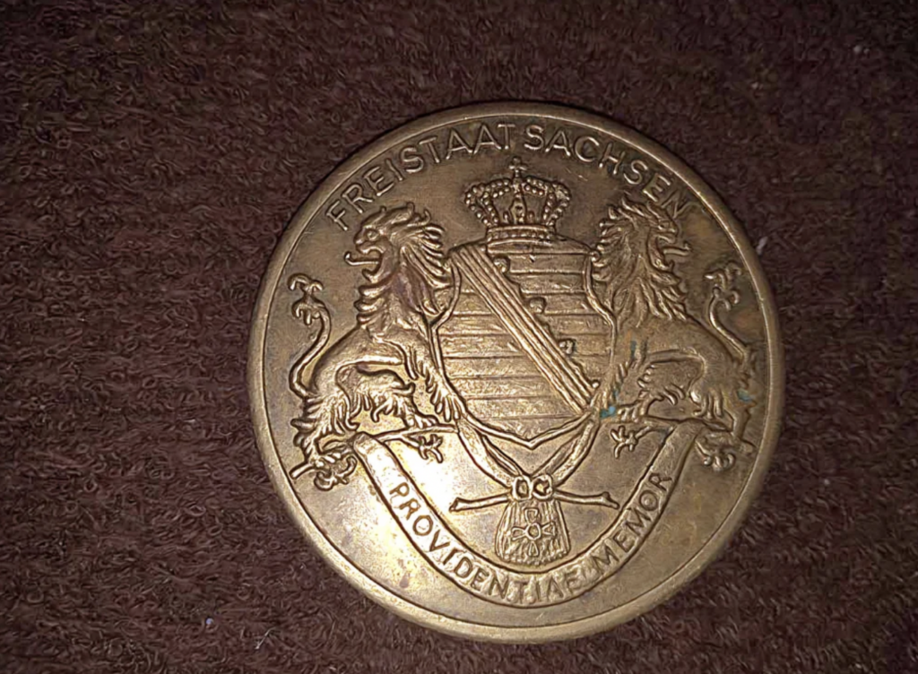 A round coin displaying the emblem of Saxony, with two lions holding a shield topped by a crown. Text around the edge reads "FREISTAAT SACHSEN" at the top and "PROVIDENTIAE MEMOR" at the bottom. The coin is placed on a textured brown surface.