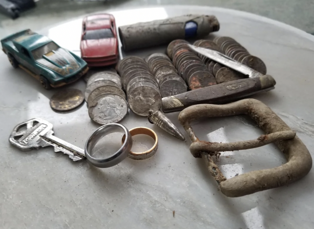 A collection of various items including coins, toy cars, a rusty penknife, keys, rings, a bullet, and a large metal buckle are arranged on a white surface. The items exhibit signs of age and wear.